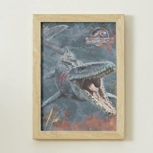 Peut inclure: Impression encadrée représentant un Mosasaure de Jurassic World. L'image montre le dinosaure avec la gueule ouverte, sur un fond sombre et texturé. Le cadre est en bois clair.