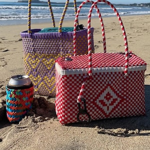 May include: Two woven baskets with handles, one purple and yellow, the other red and white, on a sandy beach. A colorful can cooler is in the foreground.