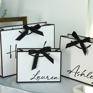 May include: Three white gift bags with black trim and black ribbon bows. The bags are personalised with the names "Ashley", "Lauren", and "H".