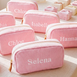 May include: A collection of rectangular, light pink corduroy pouches with gold-tone zippers. Each pouch features a white embroidered name, including Chloe, Isabella, Hanna, and Selena. Small, square gift boxes with pink ribbons are arranged nearby.