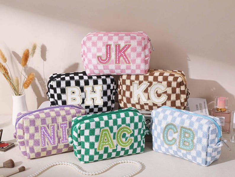 May include: Six checkered cosmetic bags in pink, black, brown, purple, green, and blue. Each bag features a different set of initials in a contrasting color. The bags are arranged on a white surface with a pearl necklace and makeup brushes.