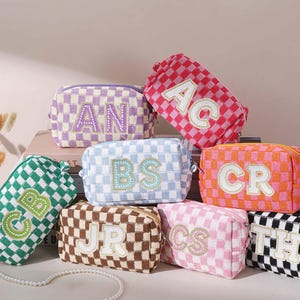 May include: A collection of personalized cosmetic bags in various colors, including pink, green, and orange. Each bag features a checkered pattern and embroidered initials. The bags are rectangular with a zipper closure.
