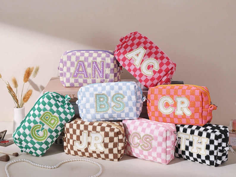 May include: A collection of personalized cosmetic bags in various colors, including pink, green, and orange. Each bag features a checkered pattern and embroidered initials. The bags are rectangular with a zipper closure.
