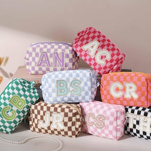 May include: A collection of personalized cosmetic bags in various colors, including pink, green, and orange. Each bag features a checkered pattern and embroidered initials. The bags are rectangular with a zipper closure.