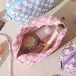 May include: A collection of cosmetic bags in various colors, including pink, blue, and purple, with a checkered pattern. One bag is open, revealing makeup brushes and products. Other items include a compact of red blush, a pearl necklace, and a perfume bottle.