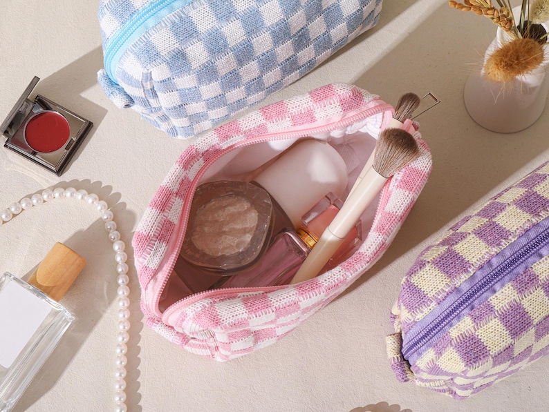 May include: A collection of cosmetic bags in various colors, including pink, blue, and purple, with a checkered pattern. One bag is open, revealing makeup brushes and products. Other items include a compact of red blush, a pearl necklace, and a perfume bottle.