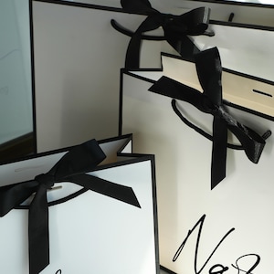 May include: Three white gift bags with black ribbon bows and black trim. The bags are personalized with the names "Lauren" and "Nastasia" in black script.