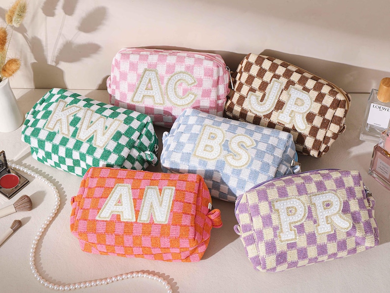 May include: A collection of six cosmetic bags in various colors, each with a checkered pattern and embroidered initials. Colors include green, pink, brown, blue, orange, and purple. The bags are arranged on a light-colored surface, with makeup and perfume bottles in the background.