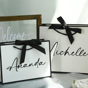 May include: Two white gift bags with black ribbon bows and personalized names. The bag on the left says "Amanda" and the bag on the right says "Michelle".