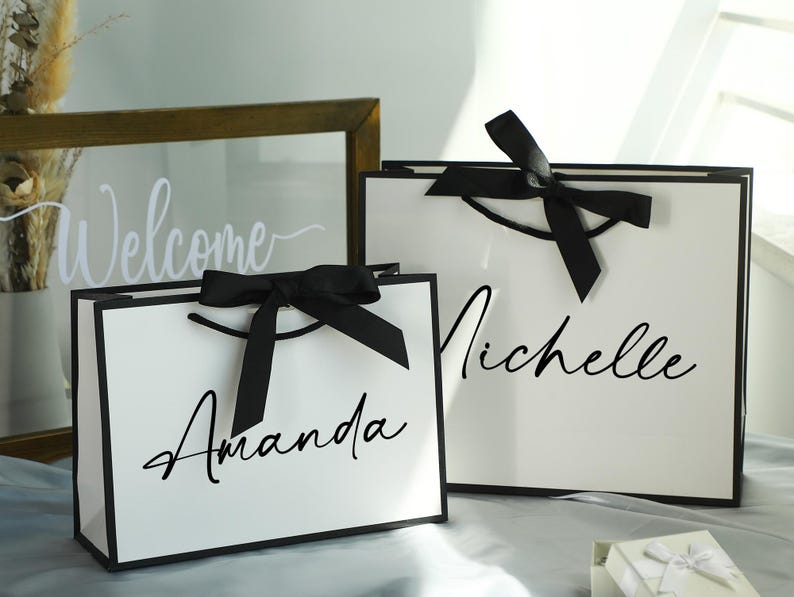 May include: Two white gift bags with black ribbon bows and personalized names. The bag on the left says "Amanda" and the bag on the right says "Michelle".