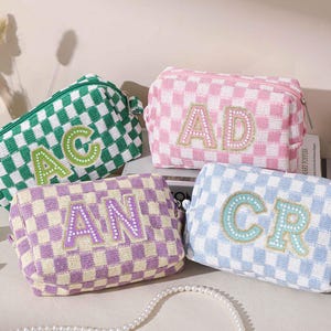 May include: Four checkered cosmetic bags in green, pink, purple, and blue. Each bag has a different set of embroidered initials. The bags are rectangular with a zipper closure. The bags are sitting on a white surface.