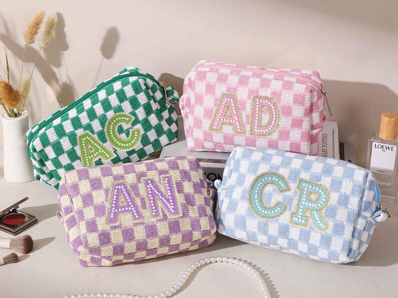 May include: Four checkered cosmetic bags in green, pink, purple, and blue. Each bag has a different set of embroidered initials. The bags are rectangular with a zipper closure. The bags are sitting on a white surface.