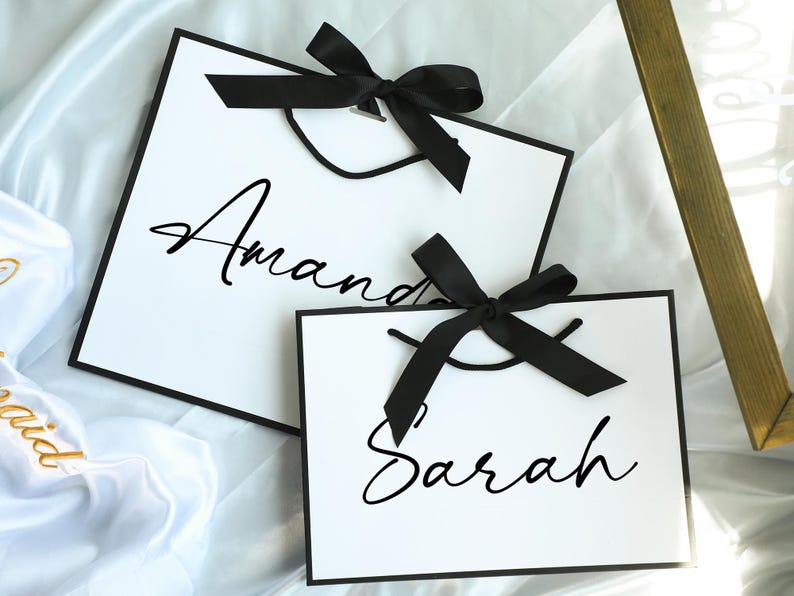 May include: Two white gift bags with black ribbon bows and black lettering. The top bag says "Amanda" and the bottom bag says "Sarah".