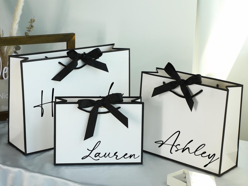 May include: Three white gift bags with black trim and black ribbon bows. The bags are personalised with the names "Ashley", "Lauren", and "H".