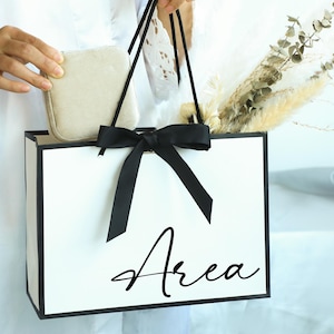 May include: A white gift bag with black trim and a black bow. The bag has the word "Area" written in black cursive on the front.