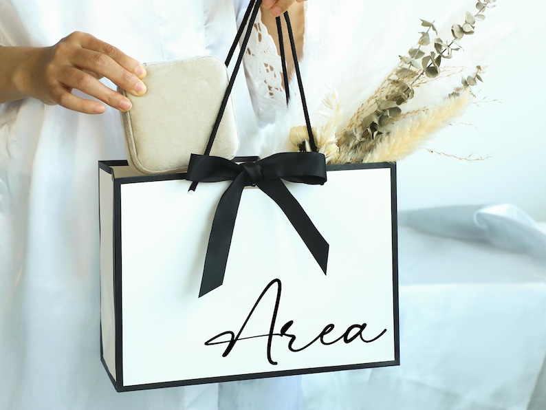 May include: A white gift bag with black trim and a black bow. The bag has the word "Area" written in black cursive on the front.