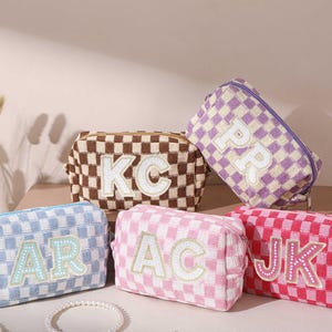 May include: Five checkered cosmetic bags in various colors: brown, purple, pink, red, and blue. Each bag features a different set of white embroidered initials. The bags have a rectangular shape with a zipper closure, perfect for storing makeup and toiletries.
