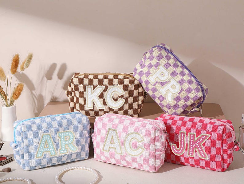 May include: Five checkered cosmetic bags in various colors: brown, purple, pink, red, and blue. Each bag features a different set of white embroidered initials. The bags have a rectangular shape with a zipper closure, perfect for storing makeup and toiletries.