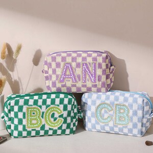 May include: Three checkered cosmetic bags in green, purple, and blue. Each bag features embroidered initials: "AN", "BC", and "CB". The bags are rectangular with a zipper closure. A perfume bottle and makeup brushes are in the background.
