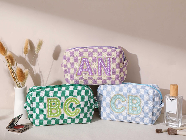 May include: Three checkered cosmetic bags in green, purple, and blue. Each bag features embroidered initials: "AN", "BC", and "CB". The bags are rectangular with a zipper closure. A perfume bottle and makeup brushes are in the background.
