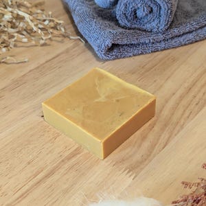 May include: A square, golden-yellow soap bar with a marbled pattern rests on a light wood surface. A rolled grey towel and a folded grey towel are in the background. Decorative dried plants are also present.