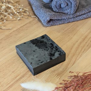 May include: A dark grey, rectangular bar of soap with a speckled design. A rolled grey towel and a folded grey towel are in the background. Dried decorative plants and wood shavings are also present.
