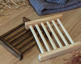 Könnte beinhalten: Zwei Seifenschalen aus Holz, eine dunkelbraune und eine natürliche, mit einem grauen Handtuch und dekorativen Elementen auf einer Holzoberfläche. Die Seifenschalen haben ein Lamellendesign zur Entwässerung. Das Handtuch ist gefaltet und gerollt.