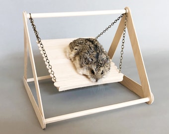 May include: A small hamster sits on a wooden swing made of popsicle sticks. The swing is suspended by a chain and has a wooden frame.