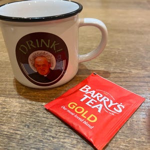 May include: A white enamel mug with a black rim and handle, featuring a circular graphic with the word "DRINK" and a portrait. A red Barry's Tea Gold tea bag is next to the mug.