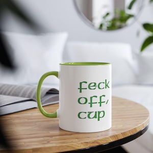 Puede incluir: Taza de cerámica blanca con interior y asa verdes, con la frase "feck off, cup" en letras verdes. La taza está sobre una mesa de madera con un fondo borroso.