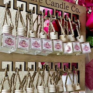 May include: A display of car air fresheners from Mi Madre Candle Co. Each small, clear glass bottle is labeled with a scent name and a floral design. The bottles hang from a wooden display with the company name at the top.