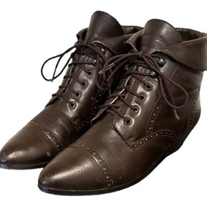 Puede incluir: Un par de botines de cuero marrón con cierre de cordones y punta puntiaguda. Los botines tienen un patrón de puntadas decorativas y un puño abatible.