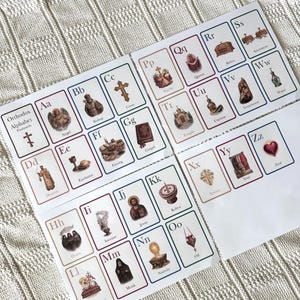 May include: Set of Orthodox Alphabet flashcards. Each card features a letter of the alphabet with an associated word and illustration. The cards are arranged on a white surface with a textured pattern. The text "Orthodox Alphabet Flashcards" is visible.