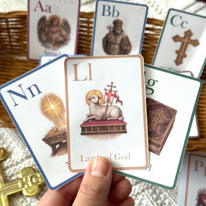 May include: A collection of illustrated flashcards featuring the alphabet. Each card displays a letter, a corresponding image, and a word. The cards are held in a hand, with a woven basket in the background. The cards have a white border and a colorful design.