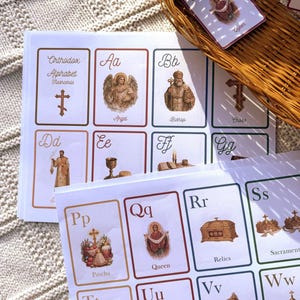 May include: A stack of Orthodox Alphabet flashcards with illustrations and text. The cards feature images of an angel, bishop, cross, and queen. A wicker basket holds additional cards, with the word "Queen" visible on one.