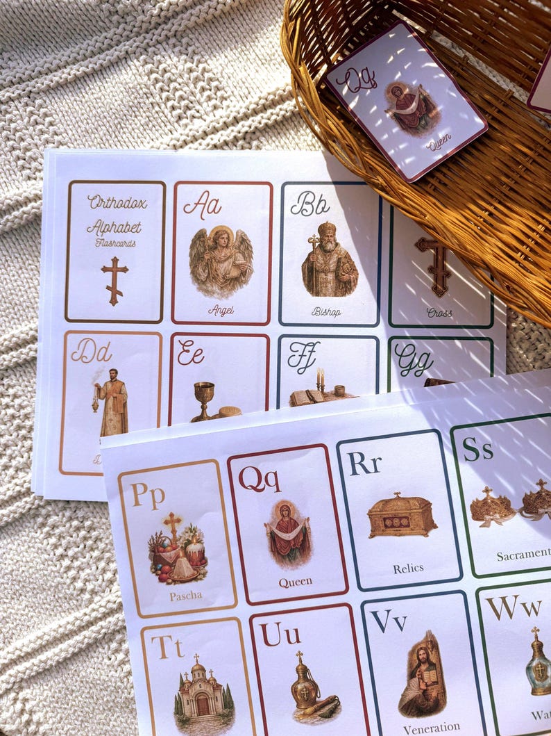 May include: A stack of Orthodox Alphabet flashcards with illustrations and text. The cards feature images of an angel, bishop, cross, and queen. A wicker basket holds additional cards, with the word "Queen" visible on one.