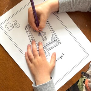 May include: A child's hands coloring a Gospel-themed coloring page with a purple pencil. The coloring page features the letter "G" and an illustration of a book with a cross on the cover. The word "Gospel" is printed on the page.