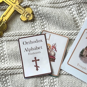 May include: A gold-colored Orthodox cross and flashcards on a cream-colored knitted surface. The flashcards are labeled "Orthodox Alphabet Flashcards" and feature a cross symbol. Other flashcards are visible with religious imagery.
