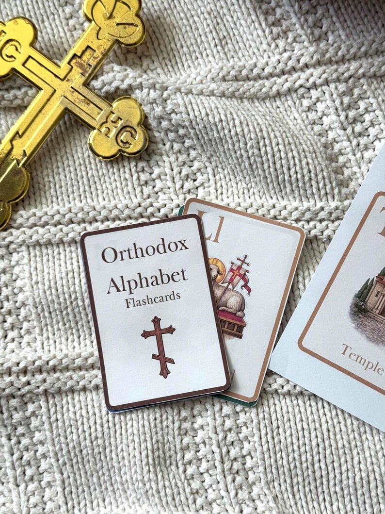 May include: A gold-colored Orthodox cross and flashcards on a cream-colored knitted surface. The flashcards are labeled "Orthodox Alphabet Flashcards" and feature a cross symbol. Other flashcards are visible with religious imagery.