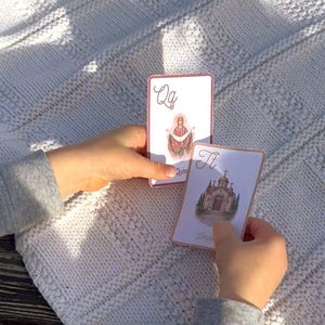 May include: Two playing cards with illustrations and text are held against a white knitted blanket. One card features the letter "Qq" and an image of a religious figure. The other card shows a building and the letter "Tt".