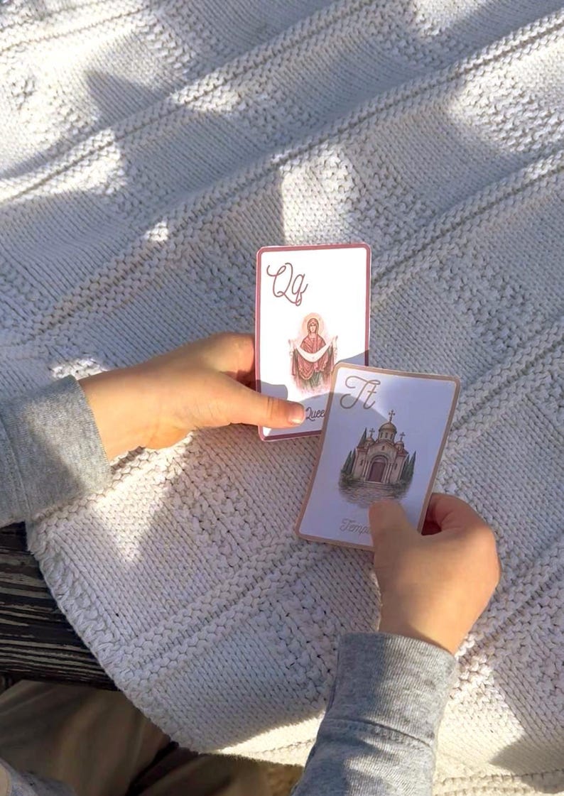 May include: Two playing cards with illustrations and text are held against a white knitted blanket. One card features the letter "Qq" and an image of a religious figure. The other card shows a building and the letter "Tt".