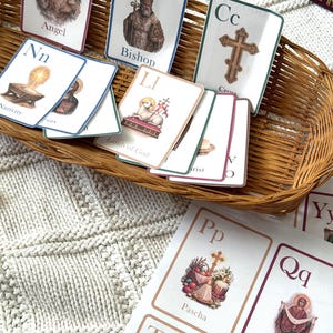 May include: A collection of educational flashcards featuring religious imagery and the alphabet. The cards are in a woven basket and on a white knitted surface. The cards include illustrations of an angel, bishop, cross, and lamb of God.