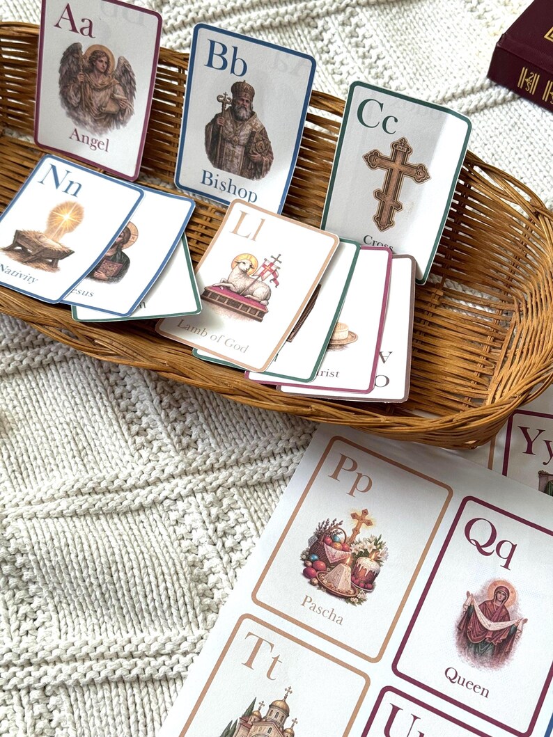 May include: A collection of educational flashcards featuring religious imagery and the alphabet. The cards are in a woven basket and on a white knitted surface. The cards include illustrations of an angel, bishop, cross, and lamb of God.