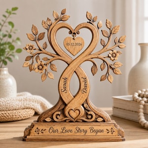 Peut inclure: Sculpture en bois en forme d'arbre avec des troncs entrelacés et un sommet en forme de cœur. La sculpture porte les noms "Sarah" et "David", un cœur avec la date "05.12.2026" et la phrase "Our Love Story Began". L'arbre a des feuilles et des fleurs.