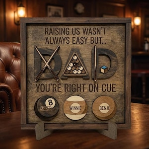 May include: Wooden sign with the word "DAD" in large letters, featuring pool cues and a rack of pool balls. The sign includes the text "RAISING US WASN'T ALWAYS EASY BUT... YOU'RE RIGHT ON CUE" and personalized pool ball designs.