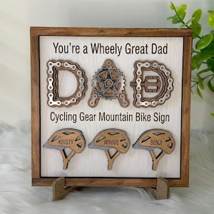 May include: Wooden sign with the text "You're a Wheely Great Dad" and "DAD" spelled out with bicycle chain and gear designs. Below are three bicycle helmets with names.