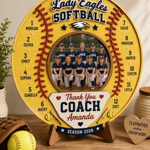 May include: A round yellow wooden softball-themed plaque featuring a central photo of a softball team, the text Lady Eagles SOFTBALL, and a list of twelve player names numbered 1 through 12. A home plate shape displays Thank You COACH Amanda.