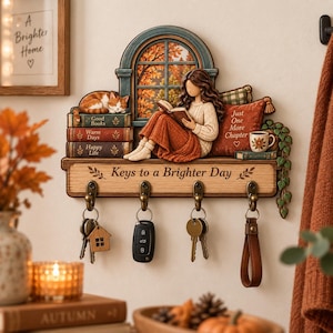 May include: A wall-mounted wooden key holder featuring a decorative scene of a person reading a book by a window with a sleeping cat on a stack of books. The shelf includes four metal hooks for keys and the text Keys to a Brighter Day.