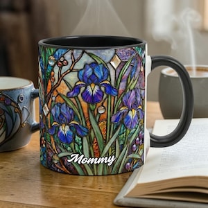 Peut inclure: Une tasse en céramique avec une anse et un bord noirs, ornée d'un motif de vitrail représentant des iris bleus et violets. Le mot "Mommy" est écrit en cursive. De la vapeur s'élève de la tasse.