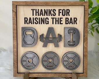 Puede incluir: Placa de madera con las palabras "THANKS FOR RAISING THE BAR" y la palabra "DAD" escrita con equipamiento de gimnasio. Incluye una pesa rusa, una barra y una mancuerna. Debajo, tres discos de pesas con nombres.
